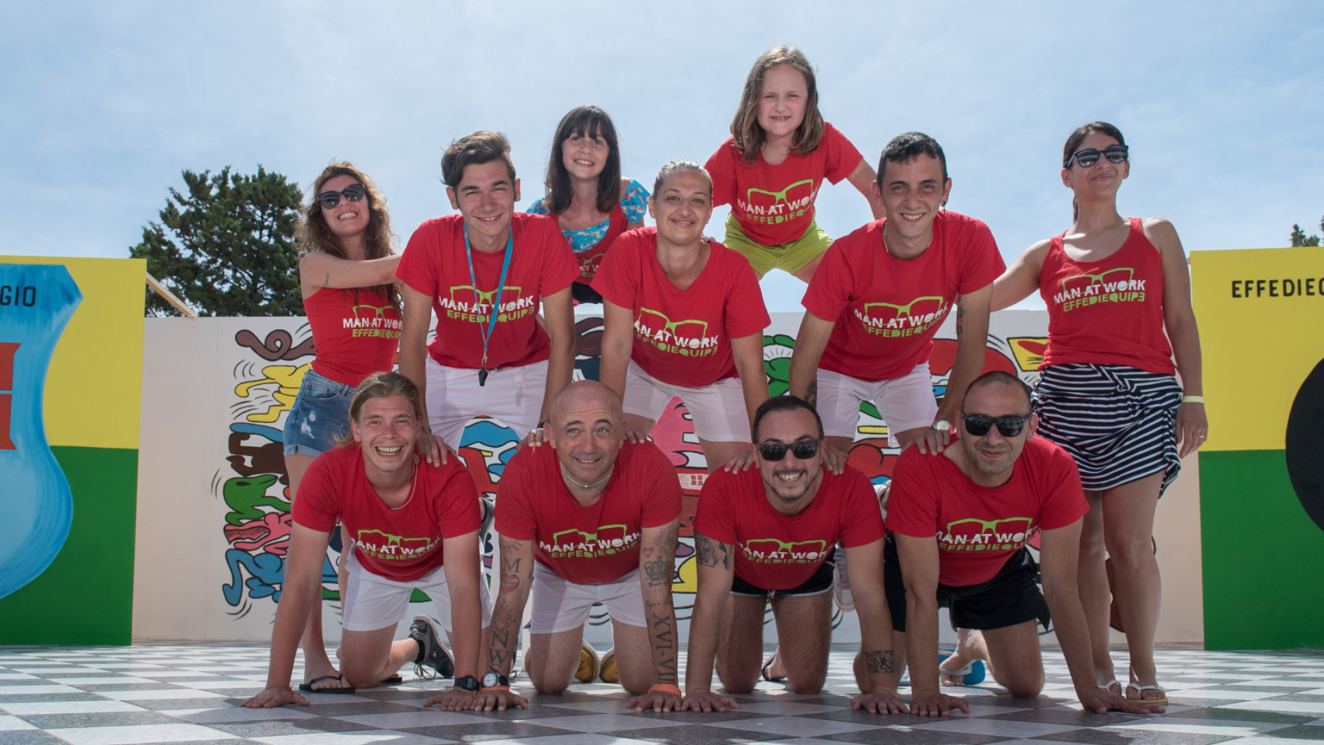 Gruppo di persone in piramide umana, indossano magliette rosse con scritta 'MAN AT WORK'.