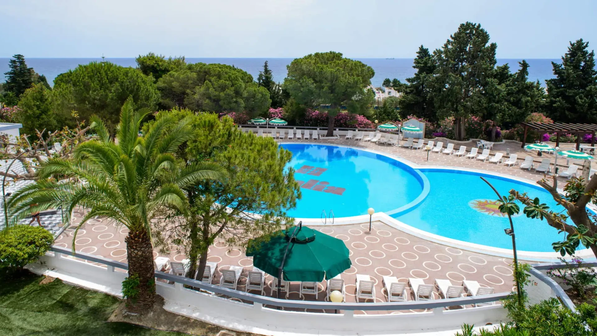 Piscina circondata da lettini, vista mare, alberi e ombrelloni verdi.