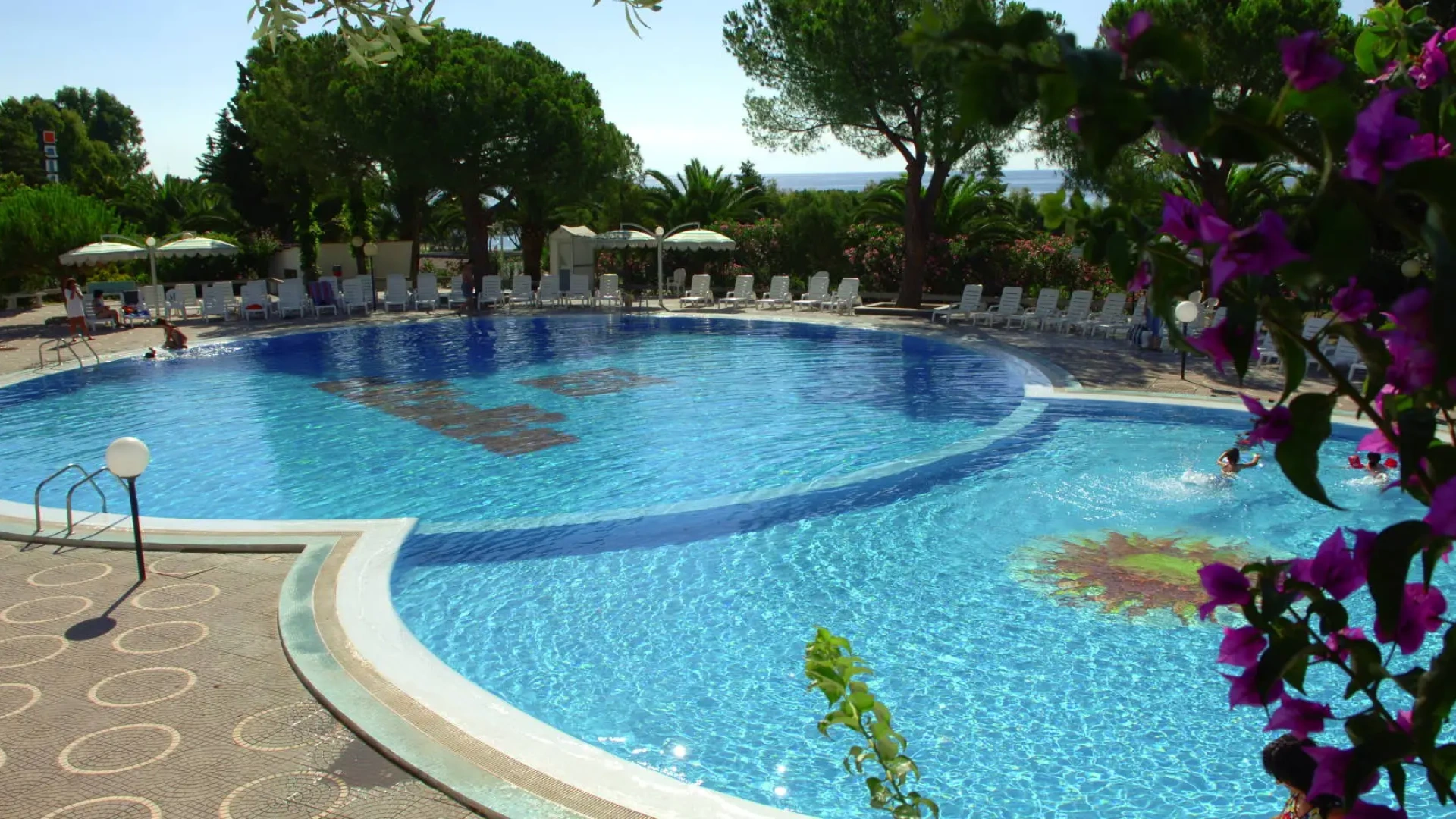 Piscina all'aperto con lettini, ombrelloni e fiori colorati intorno, immersa nel verde.