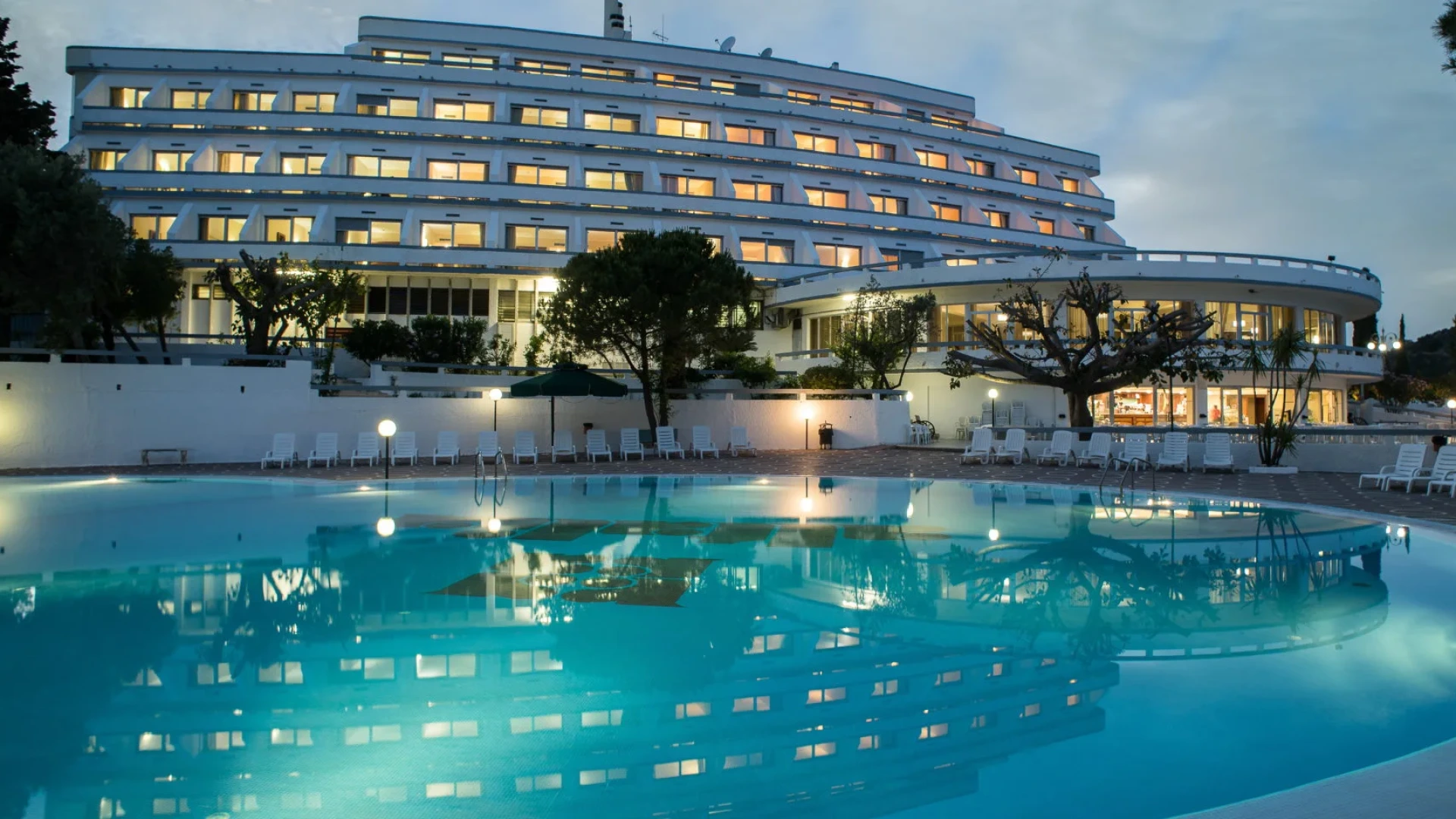 Hotel moderno con piscina illuminata e lettini, circondato da alberi e luci soffuse.