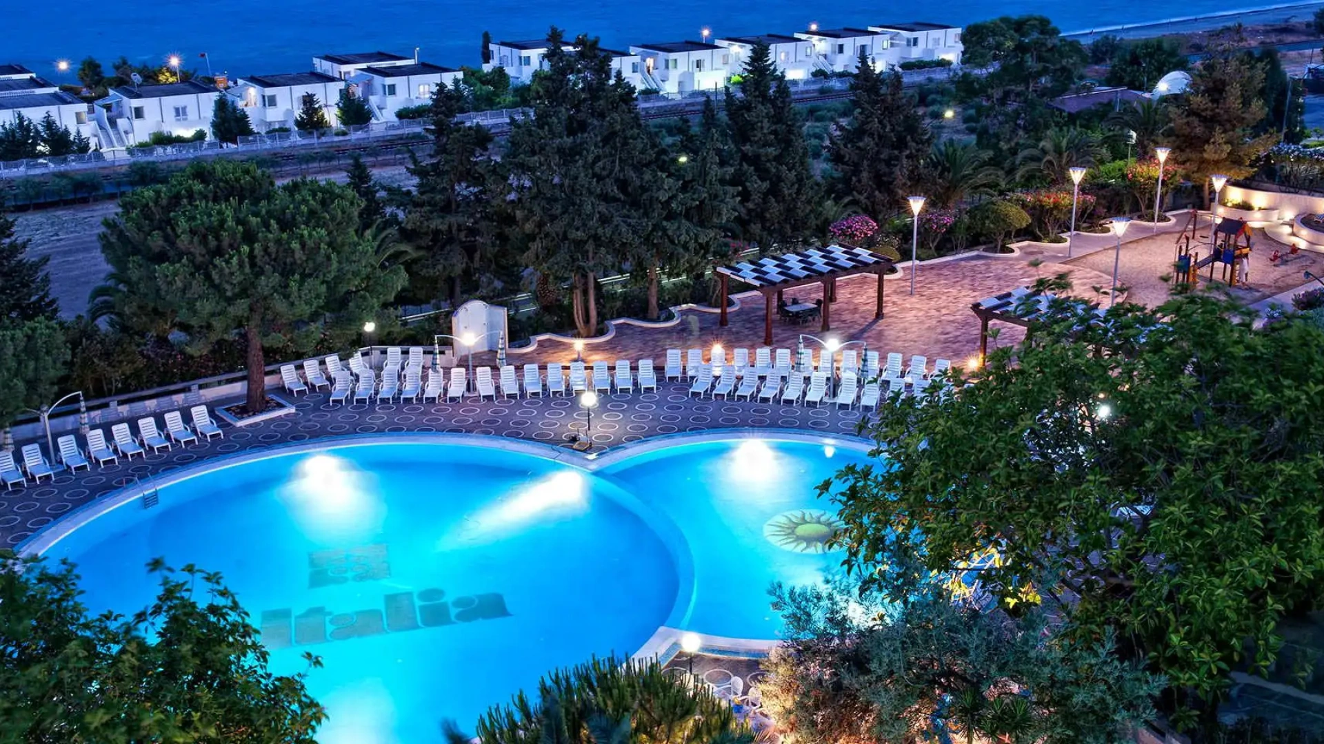 Piscina illuminata di notte con lettini, alberi e vista sul mare.