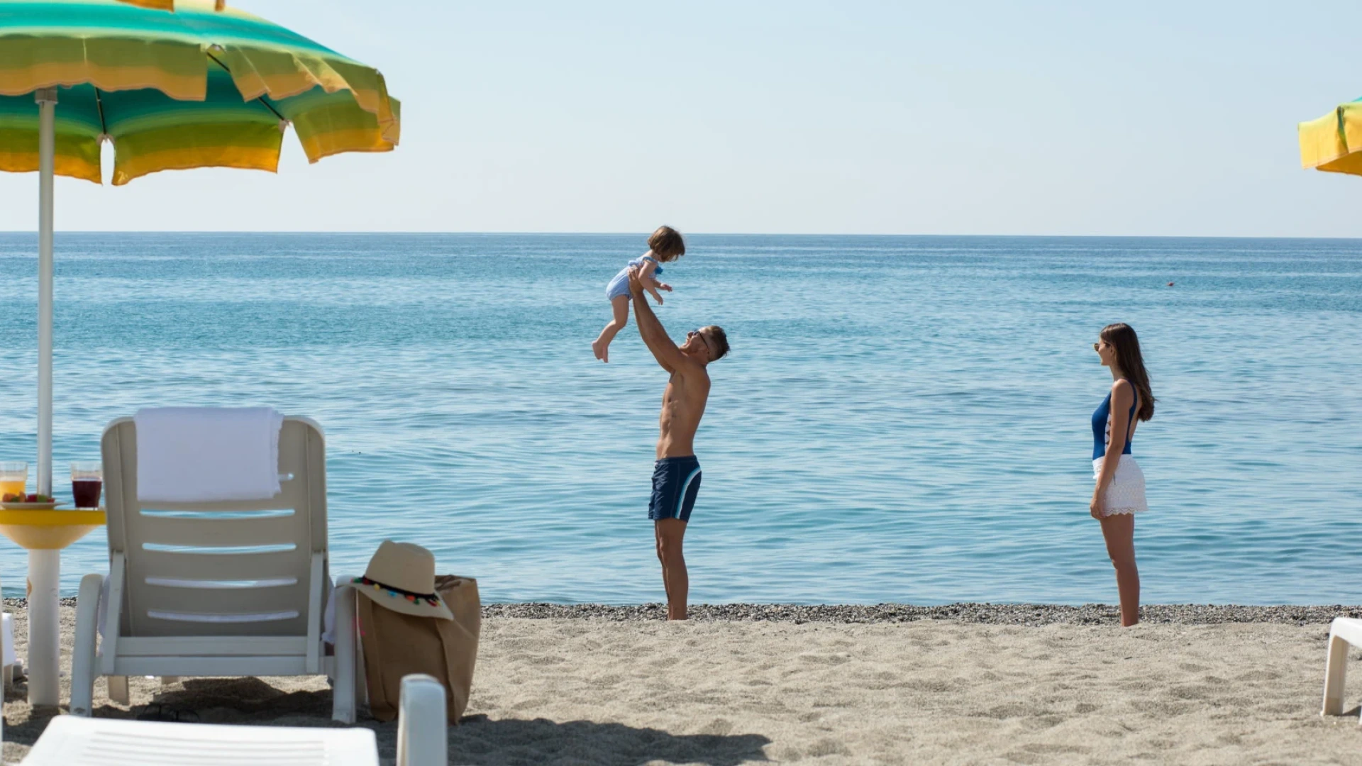 Famiglia gioca sulla spiaggia sotto un ombrellone colorato, con mare calmo sullo sfondo.